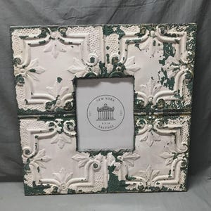 May include: Square photo frame with a distressed white and green finish. The frame has a decorative embossed pattern and a central opening for a 5x10 inch photo. The frame includes the text "NEW YORK SALVAGE".
