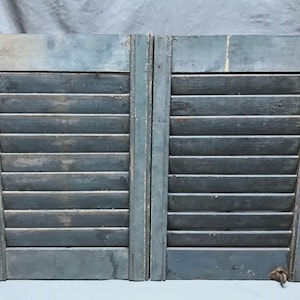 May include: Two weathered, blue-gray wooden shutters with horizontal slats. The shutters have a distressed appearance, with visible wear and tear on the paint and wood. The shutters are likely vintage or antique, adding a rustic charm.