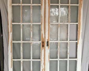 May include: A pair of white painted wooden doors with multiple glass panes. The doors have a distressed look with some of the paint chipped away, revealing the wood underneath.