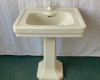 May include: Off-white pedestal sink with a square basin and a single faucet. The sink has a rectangular shape with a raised edge and sits atop a matching pedestal base. The faucet is a simple, classic design.