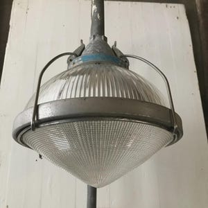 May include: An industrial-style light fixture with a clear, ribbed glass shade and a metal frame. The fixture is mounted on a metal pole and has a blue accent ring. The overall design is vintage and functional.