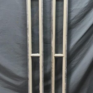 May include: Two tall, rectangular, cream-colored wooden window frames with a series of rectangular openings. The frames have a weathered appearance, suggesting they are vintage or antique. The background is a gray fabric.
