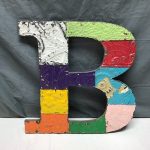 May include: A large, colorful letter B made of metal tiles. The tiles are in various colors, including white, purple, yellow, orange, green, red, blue, and pink. The tiles are attached to a wooden base with nails.