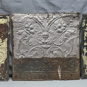 May include: Three antique, square metal ceiling tiles with ornate, embossed floral and scroll designs. The tiles are painted in shades of cream, brown, and gray, showing signs of age and wear. Each tile has a small square opening at the bottom.