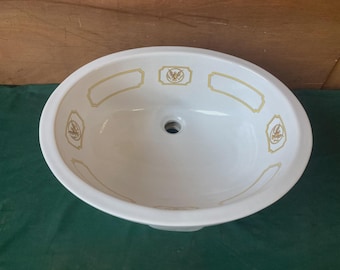 May include: An oval white ceramic bathroom sink basin featuring a central drain hole. The interior rim is decorated with a repeating pattern of gold-colored rectangular frames and a central emblem depicting an eagle with outstretched wings.