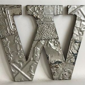 May include: A silver metal letter W made of individual pieces of tin tile. The letter is slightly distressed and has a rustic look.