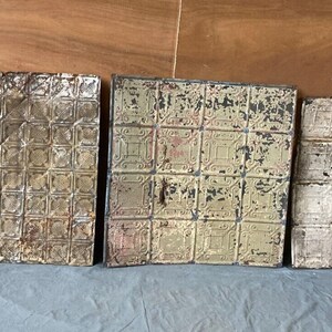 May include: Three vintage tin ceiling tiles with a geometric pattern. The tiles are in various states of wear and tear, with some showing signs of rust and paint chipping. The tiles are a light beige color with a darker beige pattern.