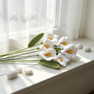 Könnte beinhalten: Ein Blumenstrauß aus fünf gehäkelten Calla-Lilien mit weißen trompetenförmigen Blütenblättern und leuchtend gelben Mitten. Jede Blume ist an einem langen, dünnen grünen Stiel befestigt, mit einem einzelnen strukturierten grünen Blatt an der Basis.