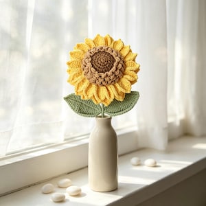 Könnte beinhalten: Eine handgefertigte Häkel-Sonnenblume mit leuchtend gelben Blütenblättern und einer strukturierten braunen Mitte, die zwei grüne Blätter an einem Stiel aufweist. Die Blume steht in einer cremefarbenen Keramik-Flaschenvase mit engem Hals.