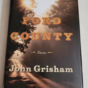 May include: A book cover with a brown background featuring a road leading into a forest with a bright sun shining through the trees. The title "Ford County Stories" is written in large, red letters with a yellow outline. The author's name, "John Grisham", is written in yellow letters below the title.