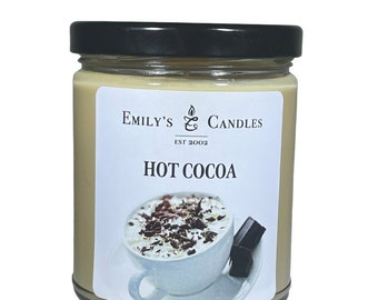 Puede incluir: Una vela de vidrio en un frasco con tapa negra y una etiqueta blanca. La etiqueta tiene el texto "Emily's Candles Est 2002" y "Hot Cocoa". La etiqueta también presenta un dibujo de una taza de chocolate caliente con crema batida y virutas de chocolate.