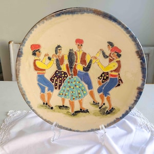 May include: A ceramic plate with a colorful illustration of six people dancing in a circle. The people are wearing traditional clothing and have red hats. The plate has a blue and white rim.