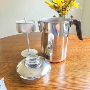 May include: A stainless steel percolator coffee pot with a black handle, a separate metal internal basket assembly with a central stem, and a circular lid featuring a clear glass knob at the top. The metal surfaces are reflective and polished.