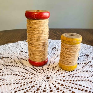 May include: Two wooden spools wrapped with twine. One spool has a red top and base, the other has a yellow top and base. The spools are sitting on a white lace doily.