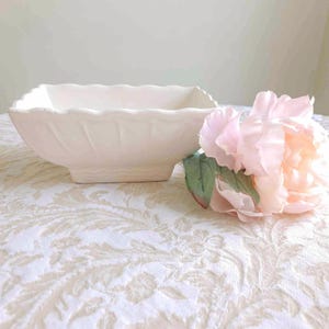 May include: A square, off-white ceramic bowl with a scalloped rim and textured sides. A pink peony flower with green leaves rests beside the bowl on a patterned tablecloth. The bowl is likely for decorative or serving purposes.