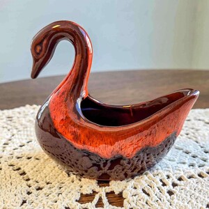 May include: A ceramic swan-shaped decorative bowl with a glossy finish. The swan features a long, curved neck and a wide, open bowl. The color scheme includes shades of brown and orange, creating a striking contrast. The bowl is likely vintage.