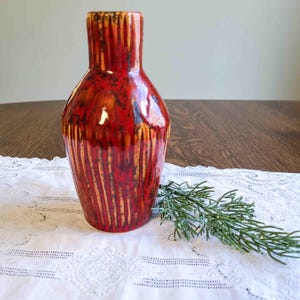May include: A glossy, red ceramic vase with vertical, textured lines and a brown rim. The vase has a rounded body and a narrow neck. It sits on a white lace tablecloth with a sprig of greenery.