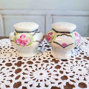 May include: A pair of vintage ceramic salt and pepper shakers. The shakers are white with a floral design featuring pink flowers and green leaves. They have a black dotted border and a white top with several holes.