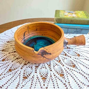 May include: A round, tan ceramic bowl with a single handle and a small pouring spout. The exterior features dark brown paint splatters, while the interior is glazed in a deep, vibrant turquoise blue. The bowl has a textured, earthy finish.