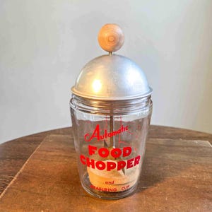 May include: Vintage glass food chopper with a metal lid and wooden handle. The words "Automatic Food Chopper and Measuring Cup" are printed in red on the glass. The chopper is sitting on a wooden surface.