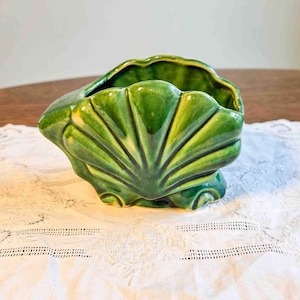 May include: A green ceramic seashell-shaped planter. The planter has a glossy finish and features detailed fan-like ridges. The interior is a darker green, and the planter sits on small, rounded feet. The planter is on a white lace tablecloth.