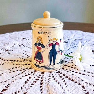 May include: A vintage mustard jar with a lid. The jar is cream-colored with a painted design of two figures and the word "MUSTARD" in black. The figures are dressed in traditional clothing. The jar is on a white lace tablecloth.