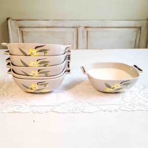 May include: A stack of six light gray ceramic bowls with yellow floral designs and brown handles. One bowl is filled with a white liquid. The bowls are arranged on a white lace tablecloth.
