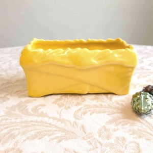 May include: A rectangular, bright yellow ceramic planter with a textured, woven-basket design. The planter has a wavy rim and is set on a patterned surface. A small, decorative green object is visible to the right.