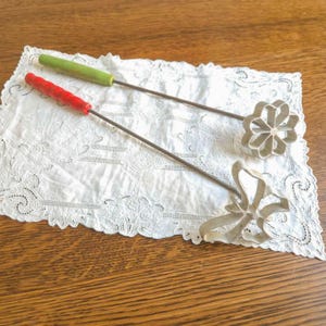 May include: Two vintage rosette irons with wooden handles. One handle is red, the other is green. The iron shapes are a flower and a bow. The rosette irons are resting on a white lace doily on a wooden surface.