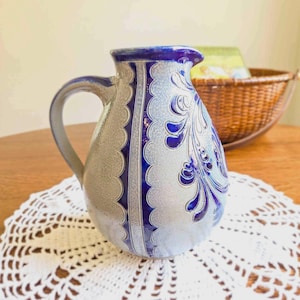May include: A ceramic pitcher featuring a light gray speckled body with a prominent blue vertical stripe and a decorative blue floral pattern. The pitcher has a curved handle, a wide spout, and a smooth, glazed finish on its rounded exterior.