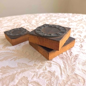 May include: Three vintage wooden printing blocks with clock face designs. The blocks are rectangular with a dark metal clock face attached to the top. The wood is a warm brown color, and the clock faces have Roman numerals.