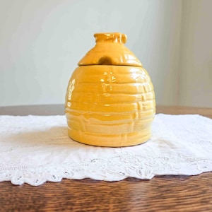 May include: A yellow ceramic honey pot with a lid. The pot has a beehive shape with horizontal ridges and a glossy finish. The lid has a small handle. The pot sits on a white lace doily on a wooden table.
