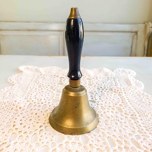 May include: A vintage brass bell with a black wooden handle. The bell has a classic design, with a flared base and a rounded top. The handle is smooth and polished, and the bell is sitting on a white lace doily.