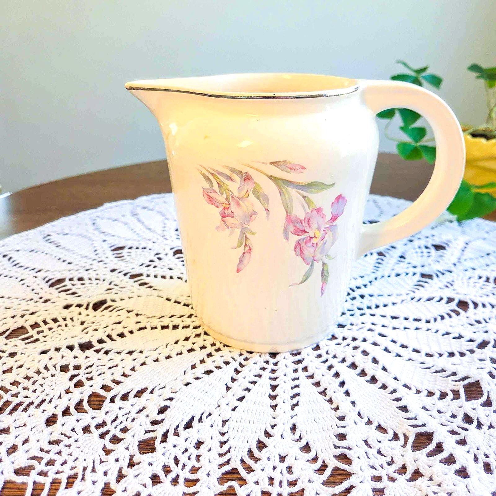 May include: Cream-colored ceramic pitcher with a curved handle and a delicate floral design in pink and green. The pitcher has a gold trim around the rim. The pitcher is sitting on a white lace doily.