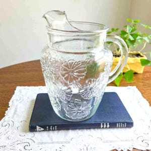May include: A clear glass pitcher with a floral pattern. The pitcher has a handle and a spout for pouring. It is sitting on a blue book and a white lace doily. A small plant in a yellow pot is in the background.