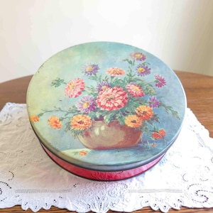 May include: Round vintage tin with a floral design. The lid features a painting of colorful flowers in a vase against a blue background. The tin has a red band around the side and sits on a white lace doily.