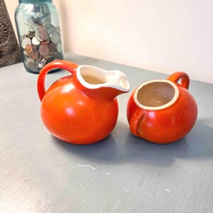 May include: A bright orange ceramic creamer pitcher with a curved handle and a white interior, paired with a matching orange ceramic sugar bowl featuring a small handle and a white interior, both showcasing a smooth, rounded, bulbous design.