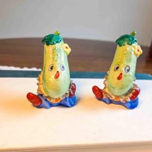 May include: Two whimsical cucumber-shaped figurines with painted faces, green tops, and blue and gold clothing. The figurines have red shoes and yellow flower accents. They are likely decorative items or collectibles.