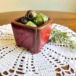 May include: A square ceramic planter with a glossy red finish and dark brown edges, filled with several small, shiny green and dark brown decorative ornaments. The planter has a textured surface and a distinct, thick rim.