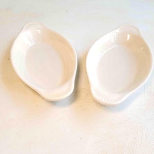 May include: Two off-white, oval-shaped ceramic dishes. Each dish has a shallow, open bowl with a slightly raised rim and a small handle on one end. The dishes are placed side by side on a white surface.