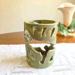 May include: A cylindrical ceramic candle holder with a matte sage green glaze. The body features large, irregular cutouts in geometric shapes, creating an openwork design that wraps around the center of the vessel, leaving solid rims at the top and base.