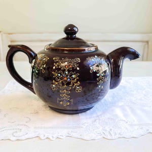 May include: A dark brown ceramic teapot with a lid, handle, and spout. The teapot is decorated with gold and colorful floral designs. The teapot is sitting on a white lace tablecloth.