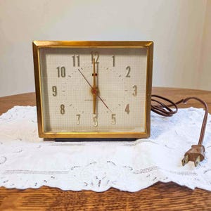 May include: A vintage General Electric alarm clock with a gold-tone rectangular frame. The clock face is off-white with black numerals and hands. The clock is plugged in with a brown cord and sits on a white lace doily.