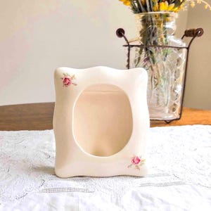 May include: A cream-colored ceramic picture frame with a smooth, wavy-edged rectangular shape. The frame features a central oval opening and is decorated with small, delicate pink rose floral motifs in the top left and bottom right corners.