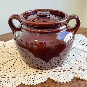 May include: A brown ceramic crock pot with a lid and two handles. The crock pot has a glossy finish and sits on a white lace doily. The lid has a small knob on top.