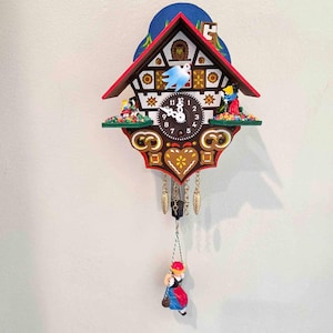 May include: A wooden cuckoo clock featuring a red roof, a central clock face with white hands, and a blue bird. The clock is decorated with painted figures, a heart motif, and two gold-colored pinecone weights. A small figure on a swing hangs below.