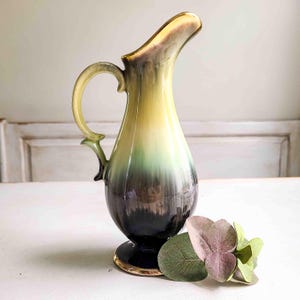 May include: A vintage ceramic pitcher with a tall, slender design. The pitcher features a gradient of colors, transitioning from yellow to green and dark gray. It has a curved handle and a gold rim at the base and spout. A decorative floral arrangement rests near the base.