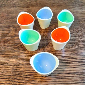 May include: A collection of six small, white, cone-shaped cups with colorful contents. The cups are filled with orange, blue, and green liquids. The cups are arranged on a wooden surface.
