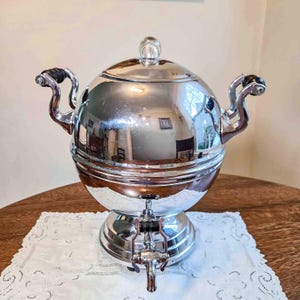 May include: A polished chrome beverage dispenser with a spherical body, a domed lid featuring a clear glass knob, and two ornate side handles with black accents. The unit stands on a circular base and includes a front-mounted spigot with a black lever.