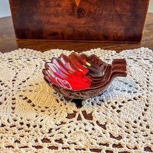 May include: A small, leaf-shaped ashtray with a rich, glossy glaze. The interior features a vibrant blend of red, brown, and black hues. The ashtray has a small, built-in rest for a cigarette and is set on a cream-colored, crocheted doily.